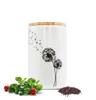 A white ceramic storage jar with a dandelion design and a bamboo lid, placed on a white background with some basil and cherry tomatoes next to it, and a pile of peppercorns. Produktbild-Vorschau