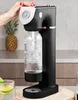 A person's hand is shown operating a black and rose gold soda maker with a bottle of carbonated water inserted. A pineapple and a glass of water with lime slices are visible in the background. Náhled obrázku produktu