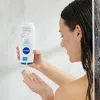 A woman is in a shower, holding a bottle of Nivea Creme Soft shower gel and dispensing it into her hand. Produktbild-Vorschau