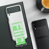 A white phone case with a green paper strip is placed on a dark book next to a black smartphone and a cup of coffee. Produktbild-Vorschau