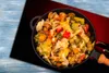 A close-up of a pan filled with stir-fried vegetables, including cabbage, peppers, and carrots, placed on a black surface with a blue and red background. Náhled obrázku produktu