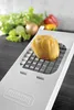 A potato is being cut on a white vegetable slicer with a grid of blades. In the background, there are fresh vegetables like lettuce, cucumber, onion, and bell peppers. Náhled obrázku produktu