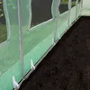 A close-up view of the bottom edge of a green greenhouse structure, showing the mesh material and metal poles, with dark soil in the foreground. Produktbild-Vorschau