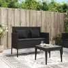 A black poly rattan garden bench with cushions and a coffee table are set up on a patio with a wooden fence and greenery in the background. Náhled obrázku produktu