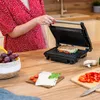 A person is using a panini grill in a kitchen setting, with ingredients and fruit nearby. Produktbild-Vorschau