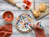 A colorful floral patterned dinnerware set is laid out on a rustic wooden table, accompanied by baked goods and flowers. Náhľad obrázku produktu