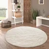 A round, fluffy cream rug is placed on a wooden floor in a living room setting, next to a ladder shelf and a plant. Produktbild-Vorschau