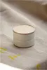 A small, round, white storage container with a light wood lid sits on a textured, light-colored fabric surface with faint yellow markings. Anteprima dell’immagine prodotto