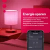 A bedside lamp is shown on a nightstand, with a smartphone displaying a smart home app interface in the foreground. The background is a bedroom setting with pink hues. Produktbild-Vorschau