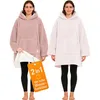 Two women wearing oversized hoodie blankets, one in pink and one in light pink, standing against a white background. A tag is attached to the pink hoodie. Produktbild-Vorschau