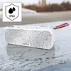 A white Bluetooth speaker with a geometric pattern sits on a wet, reflective surface, with a blurred background of trees and water. Anteprima dell’immagine prodotto