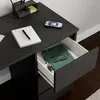 A close-up view of a black desk with a notebook, pen, and smartphone on top, and an open drawer revealing a green book and other items. Náhled obrázku produktu