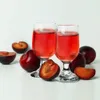 Two glasses of red liquid with plums and plum slices arranged around them on a white surface. Podgląd zdjęcia produktu