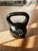 A black kettlebell rests on a wooden floor, with sunlight casting shadows. Produktbild-Vorschau