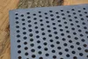 A close-up of a perforated metal grate resting on a wooden surface. Voir les images du produit