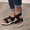 Schwarze Sandalen mit geknoteten Riemen und weißen Sohlen, getragen mit umgeschlagenen blauen Jeans auf hellem Holzboden Anteprima dell’immagine prodotto