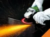 Hands wearing white gloves are using a silver and green angle grinder to cut metal, creating a shower of sparks. Produktbild-Vorschau
