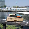 A toy pop-pop boat sits on its cardboard packaging on a railing, with a body of water and boats in the background. Produktbild-Vorschau