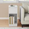 A white bedside table with books on the shelf and a cup and phone on top, next to a cream-colored sofa. Produktbild-Vorschau