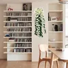 Weiße und grüne künstliche Hängeblumen mit kleinen weißen Blüten und herabhängenden grünen Blättern, präsentiert vor einer hellen Wand in einem Raum mit Bücherregalen und Holzmöbeln Náhled obrázku produktu
