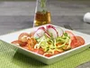 A salad is presented on a white plate, which is placed on a green placemat. In the background, a glass bottle with liquid and a fork are visible. Produktbild-Vorschau
