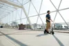 A man riding an electric scooter on a pedestrian bridge with a city skyline in the background. Produktbild-Vorschau