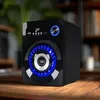 A black multimedia speaker with blue LED lights sits on a wooden table, with a blurred background of bookshelves and a plant. Produktbild-Vorschau