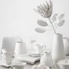 A white vase with a dried flower and other white ceramic tableware are arranged on a white surface. Produktbild-Vorschau