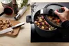 A hand is shown adjusting the lid of a pressure cooker filled with food, with ingredients and kitchen utensils laid out on a wooden surface. Produktbild-Vorschau