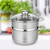 A stainless steel steamer pot with a glass lid is shown in the foreground, with a blurred background of kitchen greenery and utensils. Produktbild-Vorschau