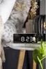 A digital alarm clock is placed on a bedside table next to a bed. Podgląd zdjęcia produktu