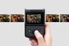A hand holding a black digital camera with a flip screen, displaying a photo of two people. Several other photos are displayed in a row to the left and right of the camera screen, suggesting a feature for reviewing or editing photos. Podgląd zdjęcia produktu