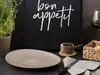 A table setting with a plate, cutlery, and a basket of bread, with a chalkboard in the background that says "bon appetit". Produktbild-Vorschau