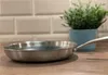 A stainless steel frying pan is placed on a wooden countertop in front of a tiled wall and a potted plant. Produktbild-Vorschau