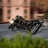 A detailed shot of a motorcycle-shaped keychain with a rider, placed on a road with blurred buildings and greenery in the background. Produktbild-Vorschau