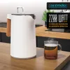 A white electric kettle with a wood-look handle sits on a wooden countertop next to a steaming mug of tea. The kitchen counter is visible in the background, along with cabinets and a faucet. Náhľad obrázku produktu