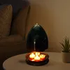 A tealight heater with lit tealights on a table next to a chair and a plant. Produktbild-Vorschau