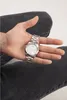 A person's hand holding a silver watch, with the person wearing dark jeans. Náhled obrázku produktu