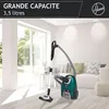 A green and gray Hoover vacuum cleaner is shown in a bright, modern living room with large windows and white walls. A sofa with decorative pillows is visible in the background. Produktbild-Vorschau