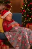 A young girl wearing a Santa hat sleeps on a couch, covered by a Christmas-themed blanket. A decorated Christmas tree and stocking are visible in the background. Náhled obrázku produktu