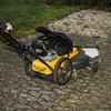 A person is pushing a yellow and black lawn mower on a paved path next to a grassy area. Produktbild-Vorschau