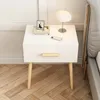 A white nightstand with a wooden drawer pull and legs, placed next to a bed. Náhľad obrázku produktu