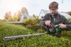 A man is using a hedge trimmer to trim a hedge in a garden. The sun is shining brightly. Produktbild-Vorschau