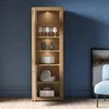 A tall, narrow wooden display cabinet with glass doors and internal lighting, showcasing various decorative items on its shelves. Produktbild-Vorschau