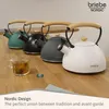 Four kettles of different colors (green, black, gray, white) are lined up on a wooden surface. A glass of tea with a tea bag is in the foreground. Produktbild-Vorschau