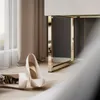 A pair of white high heels are placed on a wooden floor next to a modern furniture piece with gold accents. Produktbild-Vorschau