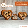 Three metal heart-shaped log holders filled with firewood, displayed outdoors on a paved surface with a wooden plank background. Produktbild-Vorschau