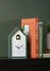 A cuckoo clock shaped like a house sits on a shelf next to books. Náhľad obrázku produktu