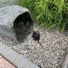 A hollow rock-like structure is placed on gravel next to a green bush. A black electrical transformer box is also on the gravel. Produktbild-Vorschau