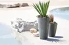 Two gray planters with wavy vertical patterns are placed on a sandy area next to a pool of water. Large rocks and smaller pebbles are scattered around the planters. Voir les images du produit